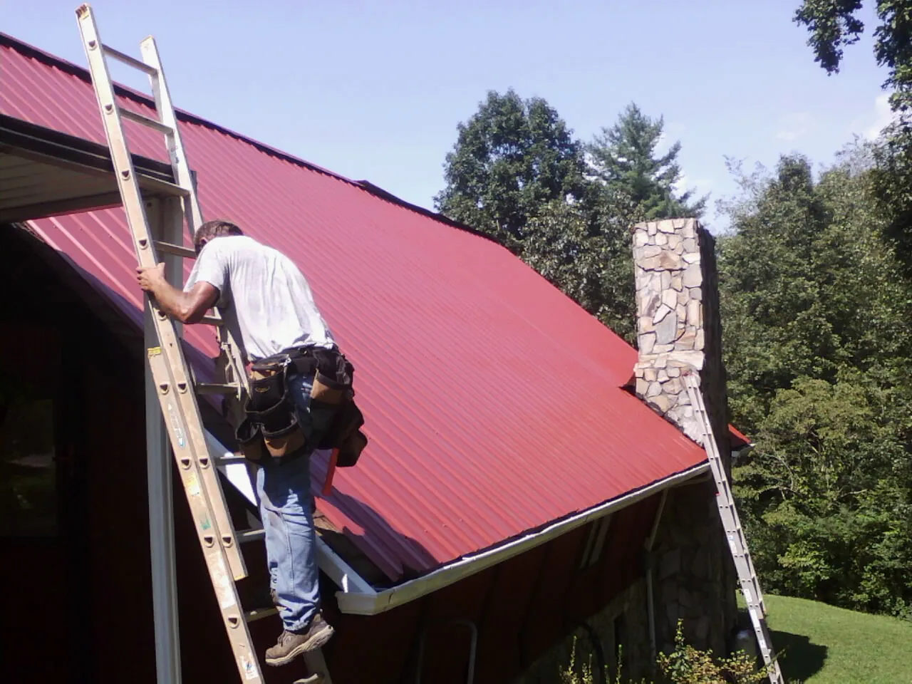 Roofer on a ladder beside a red metal roof and stone chimney — Soffit & Fascia Repair by Roofing Brothers in Walling